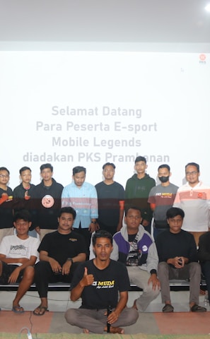 A group of people is posing for a photograph in a room with a white screen in the background that has text welcoming e-sport participants for a Mobile Legends event organized by PKS Prambanan. The group consists of 20 individuals standing and sitting, with some wearing casual attire and others wearing matching shirts that include logos.