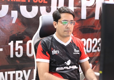 A man with glasses is sitting in a gaming chair, focused on a monitor in front of him. He is wearing a black and red esports jersey with various logos. The background features a large banner with text and numbers in bold fonts.